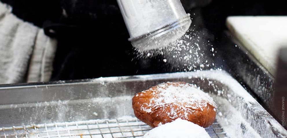 A golden beignet on a sheet tray being dusted with a heavy snow of powdered sugar at Brenda's French Soul Food.