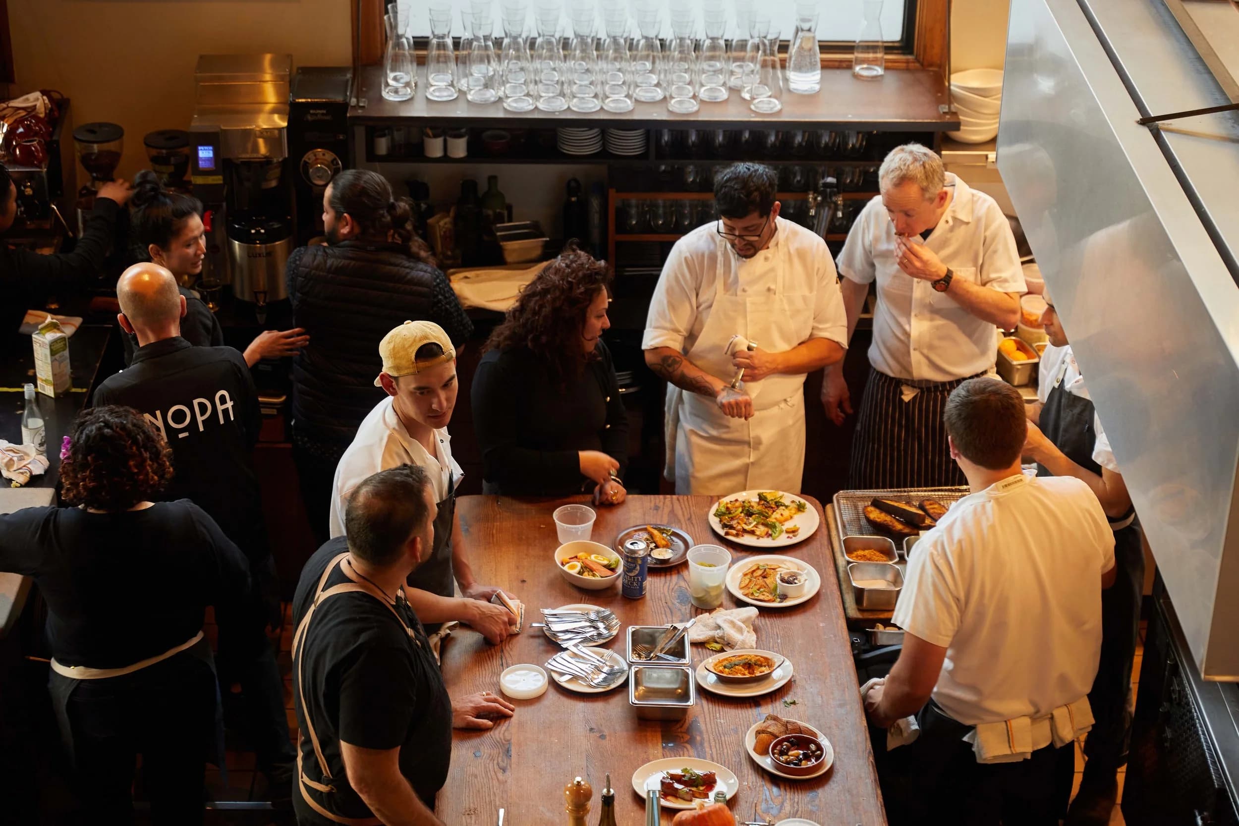 An overhead view of Nopa's busy chef's counter, staff plating dishes from the daily wood-fired menu